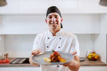 Portrait of Caucasian chef in uniform holding plate with salmon and orange fruit. Selective focus on chef.