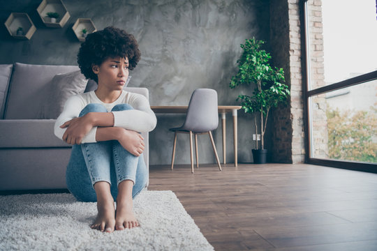 Photo of pretty dark skin curly lady sitting floor near sofa sad missing boyfriend looking window offended wear casual sweater jeans outfit flat indoors