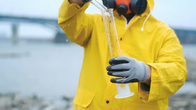 biotech expert in a respirator pours water from the river into a test tube for bacteriological analysis. Carefully examines the contents. Shakes to determine contamination. Ecology is modern.