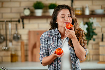 Young woman in kitchen. Beautiful woman smelling tomato. 