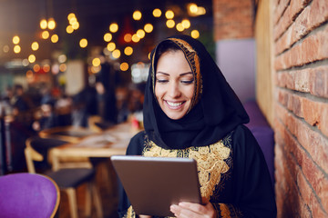 Gorgeous muslim woman dressed in traditional wear sitting in cafe and using tablet.