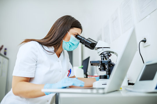 Charming caucasian laboratory assistant in white uniform, with protective mask and rubber gloves sitting in lab looking blood sample trough microscope and using laptop.