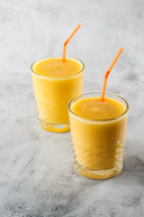 Two glasses of yellow banana, orange , mango smoothies or juice fruit isolated on bright marble background. Overhead view, copy space. Advertising for cafe menu. Vertical photo.