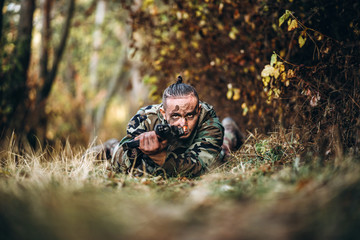 Portrait of a camouflage soldier with rifle and painted face lying in the grass aiming at the rifle. Playing airsoft outdoors in the forest