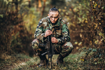 Portrait of a camouflage soldier with rifle and painted face sitting in the grass. Airsoft outdoors in the forest