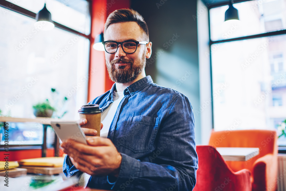 Poster half length portrait of successful bearded male blogger with tasty coffee in hands smiling at camera