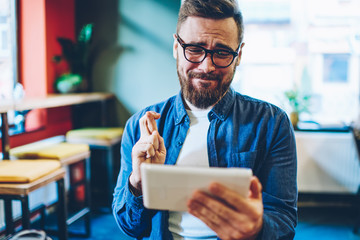 Emotional young man with crossing fingers watching online broadcasting on modern touch pad...