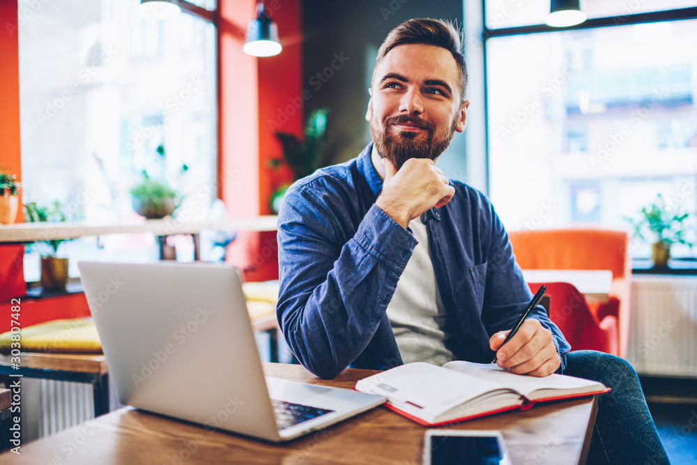 Poster smiling young bearded man writing plans in notebook and dreaming on business project sitting at netb