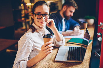 Half length portrait of prosperous student holding tasty coffee in hands and smiling at camera while collaborating with male partner sitting at modern computer with wireless 4G internet in coworking