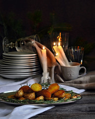 food photography of christmas baked spicy potato with rosemary on a metal plate side view on the burning candles, holiday glassware and dark texture background and old wooden table close up