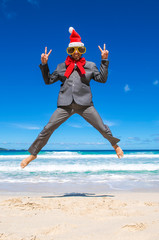 Happy businessman wearing Santa hat, sunglasses and large red Christmas bow jumping for joy on a tropical beach