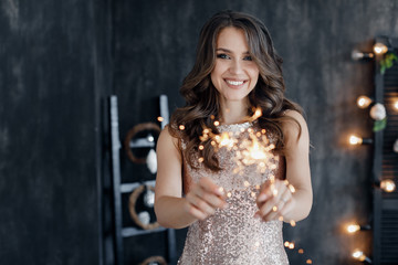 Girl with a sparkler near the Christmas tree. A cheerful young woman with a cute smile in a beige dress is standing and holding a sparkling sparkler in the hands against the background of the Christma - 303812879