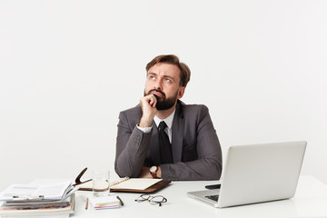 Puzzled young bearded brunette man with short haircut wearing formal clothes and wristlet watch while sitting at table with modern laptop and working notes over white background