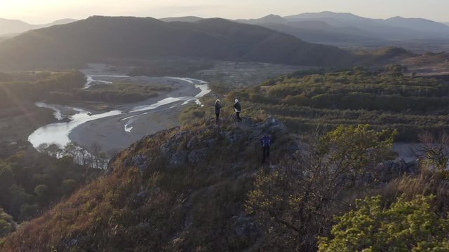 Aerial shot of  people standing  on top of a huge cliff with the view on valley with riverbed taking a one hundred eighty degree turn and mountain ridge  in the background on the sunset.