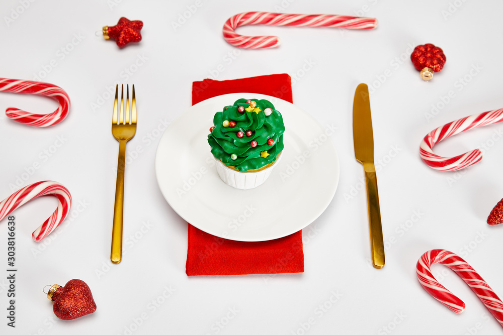 Wall mural delicious cupcake on white plate with golden cutlery, candies, baubles and red napkin on white surfa