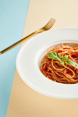 selective focus of delicious spaghetti with tomato sauce and basil in plate near fork on blue and yellow background