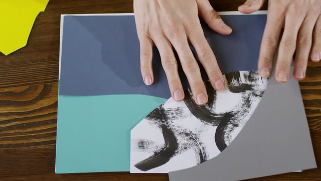 Top view close up shot of hands of unrecognizable woman removing colored and patterned paper pieces from collage background, leaving only a sheet of white paper on desk