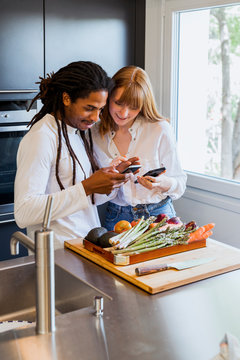 Vertical photo of a multicultural couple in the kitchen looking at the mobile next to a window