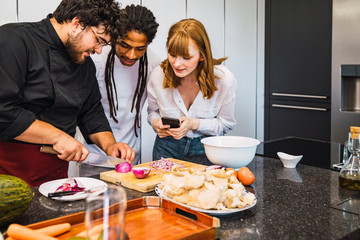 Cook cutting an onion and showing it to two young people of different ethnicities with a mobile in his hand