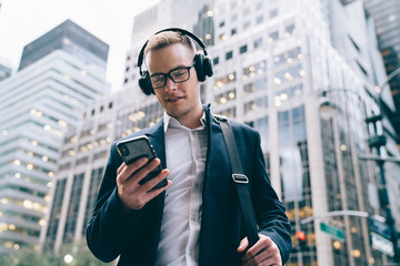 Confident elegant young man using headphone while browsing smartphone on street