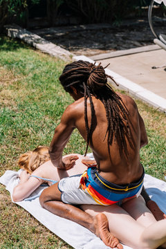 Vertical photo of a black guy with dreadlocks massaging a white girl lying on the lawn