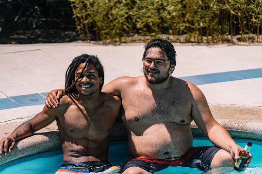 Two boys of different ethnicities sitting in a swimming pool holding on to their shoulders with a beer in their hand