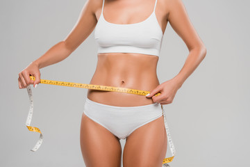 partial view of beautiful slim woman in underwear holding measuring tape on waist isolated on grey
