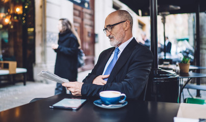 Senior bald man with grey beard reading newspaper in street cafe