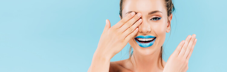 Obraz premium happy beautiful woman with blue lips covering eye with hand isolated on blue, panoramic shot