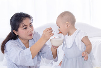 Beautiful Asian woman mother feeding fruits nutrition to her toddler girl baby by spoon, adorable baby girl eating fruits in bowl, happy motherhood relationship taking care between mother and daughter