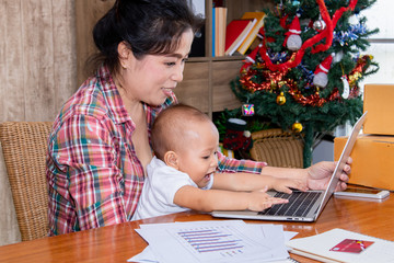 Beautiful Asian mother woman with baby working at home using laptop, smartphone checking package products, small business startup entrepreneur, freelance lifestyle shopping online holiday season