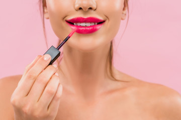 partial view of smiling naked beautiful woman with pink lips applying lip gloss isolated on pink