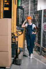 cheerful workwoman standing near forklift loader with hand on hip, touching helmet and looking at camera