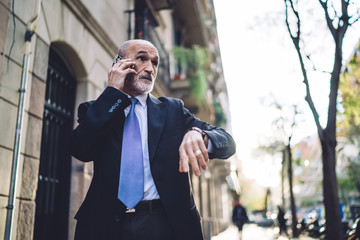 Confident bearded senior businessman in suit talking on cellphone while checking time on street