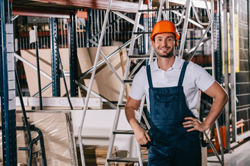 Handsome warehouse worker standing with hands on hips and smiling at camera