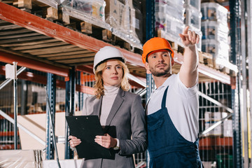 handsome warehouse worker pointing with hand while standing near businesswoman in storehouse