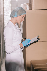 Fototapeta na wymiar focused storekeeper in hairnet writing on clipboard while standing near carton boxes in warehouse