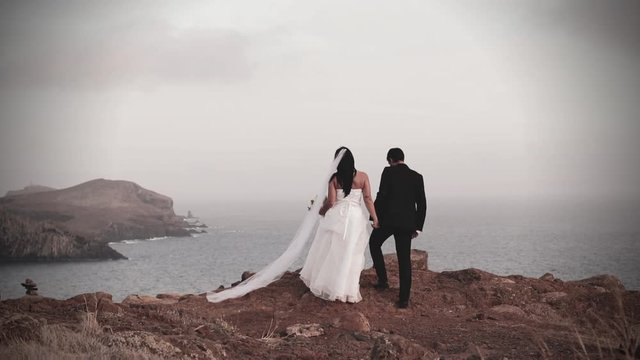 Newly married couple holding hands walking together towards arock ridge at sunset on wedding day at remote seaside coastal location celebrating love for bridal photoshoot panaromic natural color grade