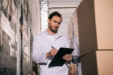 concentrated storekeeper in white coat writing on clipboard in warehouse