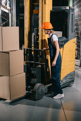 Fototapeta na wymiar workwoman in overalls and helmet standing near foklift loader in warehouse