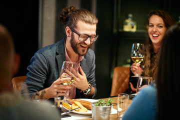 Handsome caucasian bearded hipster man with eyeglasses drinking wine and laughing while sitting with his friends in restaurant.