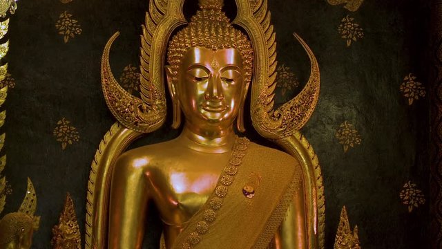 A tilting, closeup shot of the golden cast Buddha, Wat Phra Chinnarat, with a couple of cast guardian beasts image of Garuda, against a black wall background.