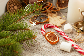 Cozy winter holidays still life on a gray knitted background with gingerbread, spruce branches, dried citruses, pumpkin, Christmas canes, burning candles and lights.