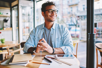 Obraz premium Handsome smiling smart male entrepreneur enjoying work break and pondering about creative ideas for business startup.Mature clever positive man in stylish eyeglasses looking away while sitting in cafe