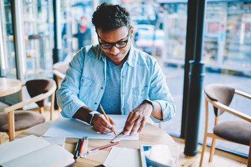 Fototapeta premium Concentrated hindu male architect drawing building blueprint sitting at desktop with equipment in coworking space.Pensive young man designer with pencil and line working on sketch in studio