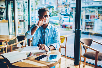 Fototapeta premium Handsome smart talented male architect in trendy eyeglasses looking away while drinking water from glass cup in cafeteria.Mature clever hindu employee enjoying working break sitting at wooden table