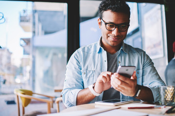 Fototapeta premium Hindu young man checking social networks and reading sms message on smartphone sitting in coffee shop.Casual dressed hipster guy in eyewear checking mail on mobile phone via 4G internet connection