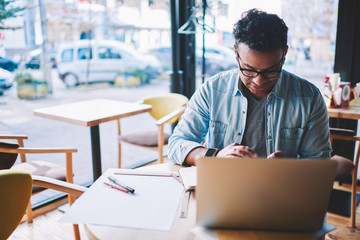 Pondering clever male architect dressed in trendy denim clothing pondering while sitting at wooden table and solving working issue .Contemplative businessman planning schedule using modern device