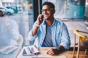 Handsome smiling male person in eyeglasses enjoying time while communicating with friends via application on smartphone.Good looking emotional man talking on modern telephone during sitting at table