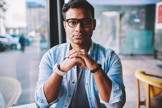Half length portrait of serious intelligent hindu businessman in glasses for eye protection pondering about creative ideas for startup.Cropped image of male person looking at camera while sitting